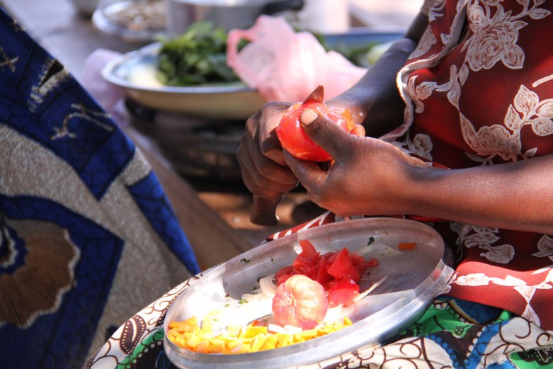A black woman peeling a tomato over a plate with a peeled tomato, peels, and chopped pumpkin, with greens in the background.
