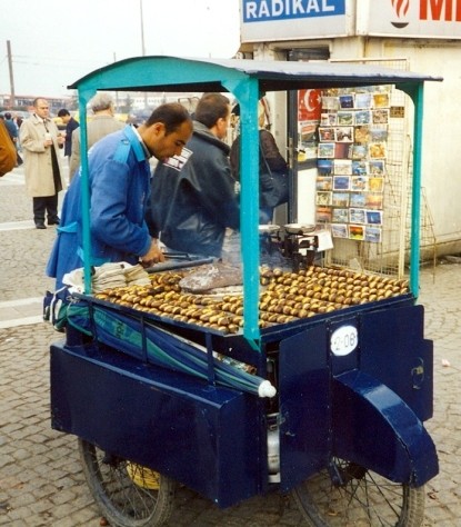 A kestaneci, or roasted chestnut vendor, in Istanbul. He is wearing a blue jacket with roasted chestnuts and a roasting pan on a blue cart.