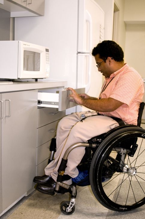 A man using a wheelchair opening a drawer under a microwave in front of a fridge.