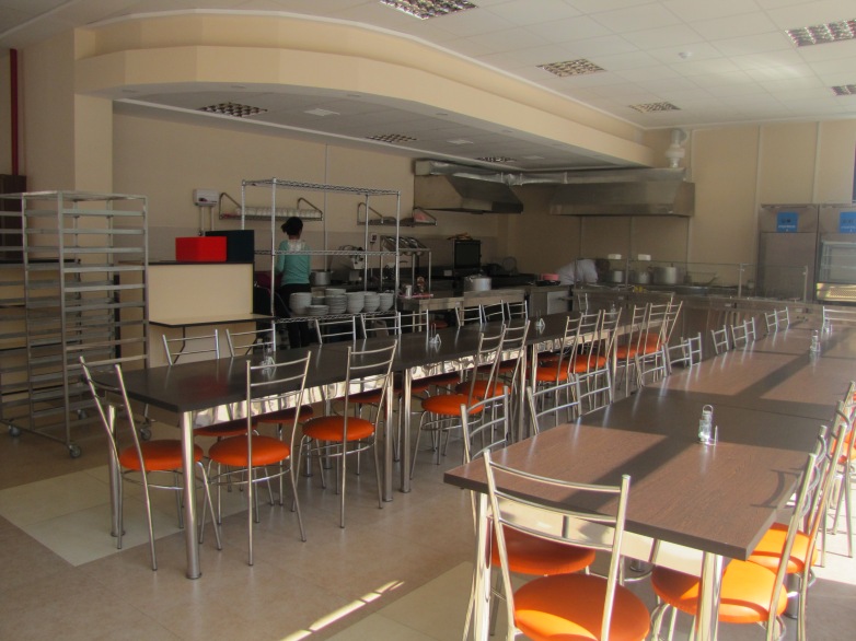 Long tables with chairs with orange seats arrayed in a cafeteria with a food service area with a stove and stacked bowls behind it. Rolling trays are on the left.