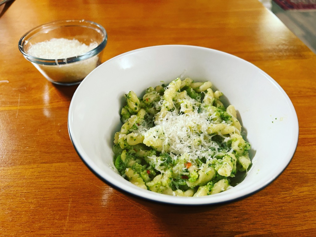 Twisted pasta with a broccoli sauce and cheese in a black and white bowl with a glass dish of grated cheese behind it. Some red pepper flakes are visible.